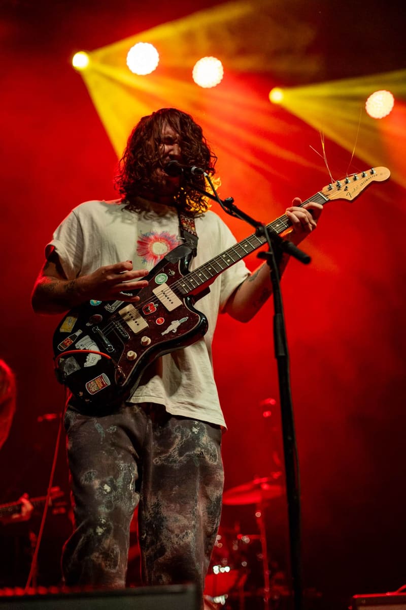 Musician with long dark hair performs on electric guitar under red and yellow stage lights.