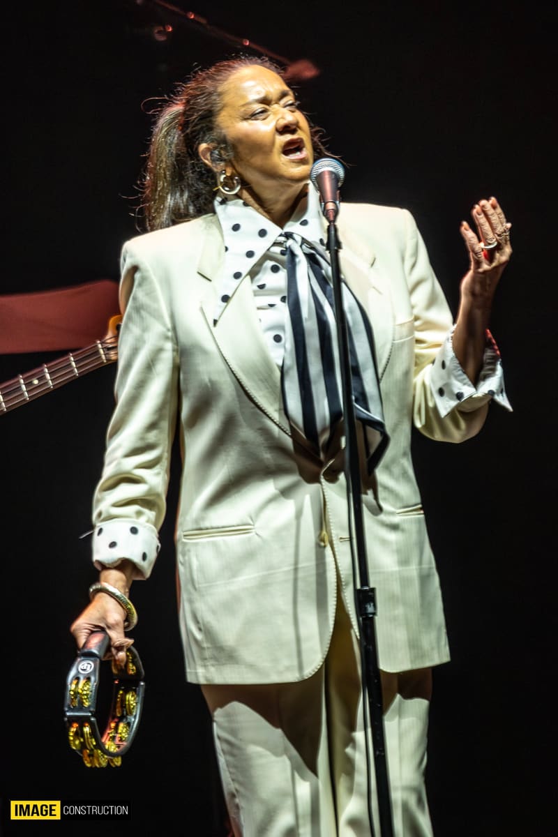 Singer in cream blazer and striped tie performing at microphone under stage lighting.