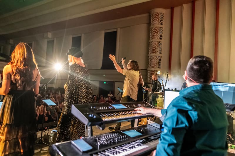 Musicians perform at keyboards and synthesisers in a gallery space with classical architectural detailing.