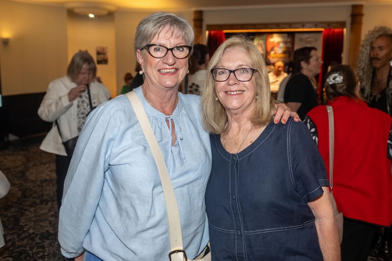 Two smiling people stand together at a gallery opening, with other visitors visible in the warmly lit background.