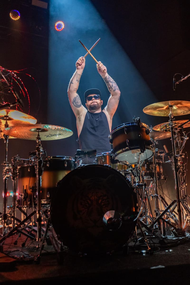 Drummer in sunglasses and black beanie holds drumsticks aloft on a lit stage, surrounded by multiple cymbals and drums.