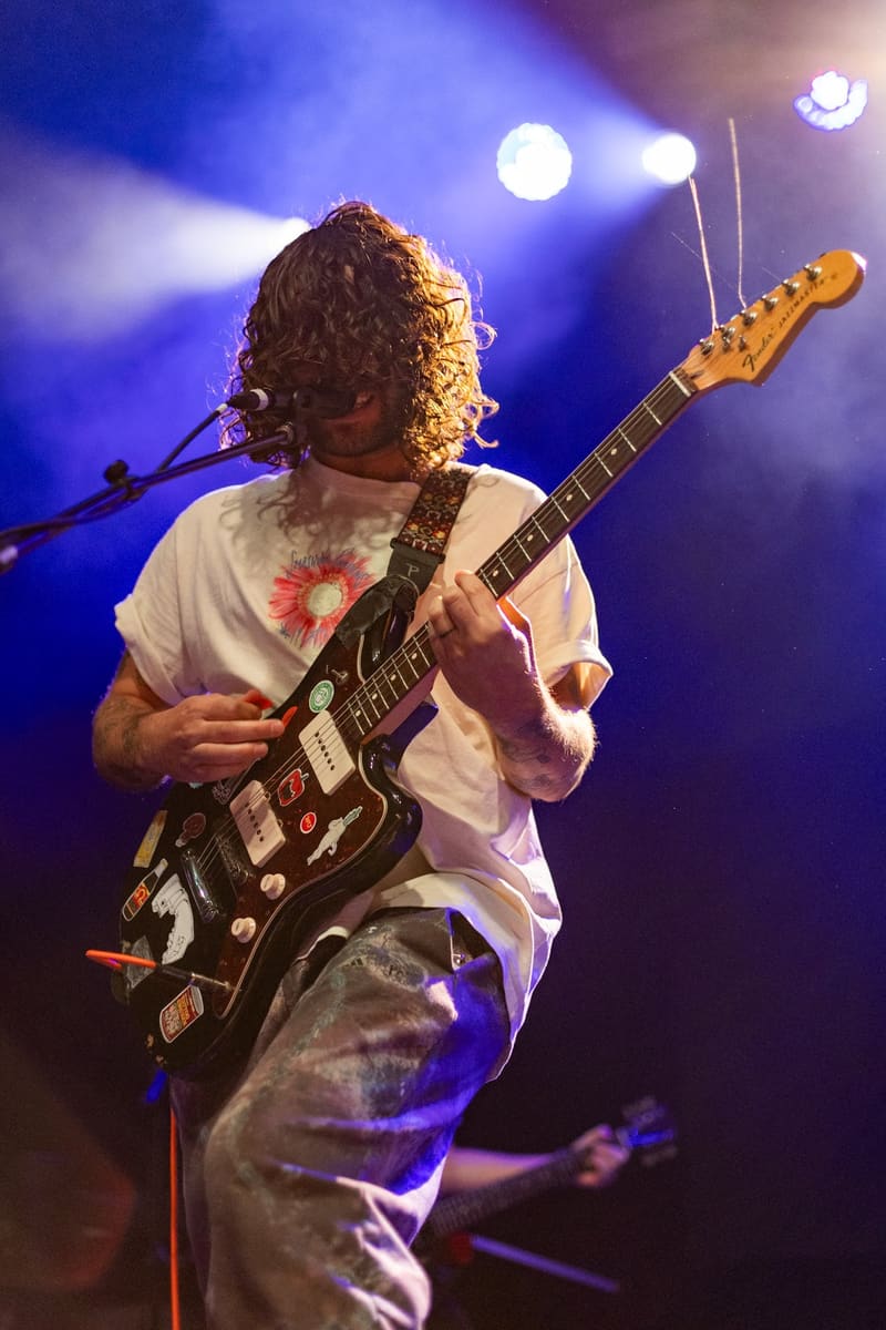 Guitarist with curly hair performing on stage under blue spotlights, holding a black electric guitar.