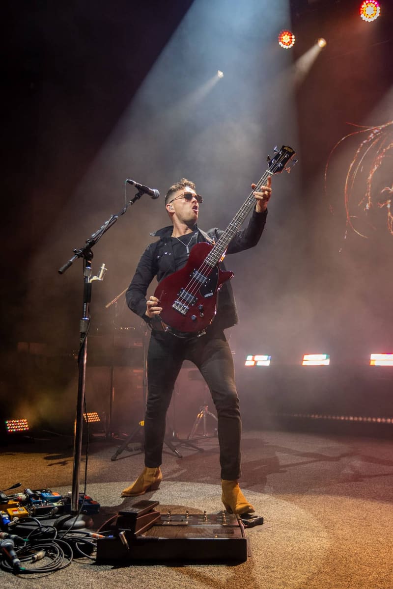 Bassist and vocalist performing on stage with microphone and bass guitar amid atmospheric stage lighting and smoke.