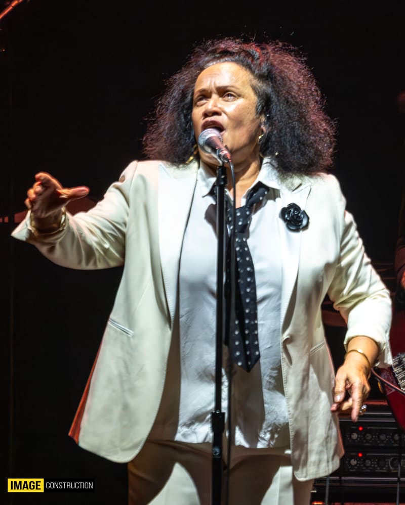 Singer performing at microphone in cream blazer and dark patterned shirt during stage concert.