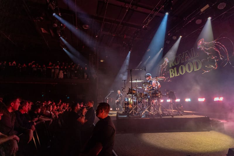 Rock band performs on stage beneath dramatic blue and red lighting with crowd silhouetted in foreground.