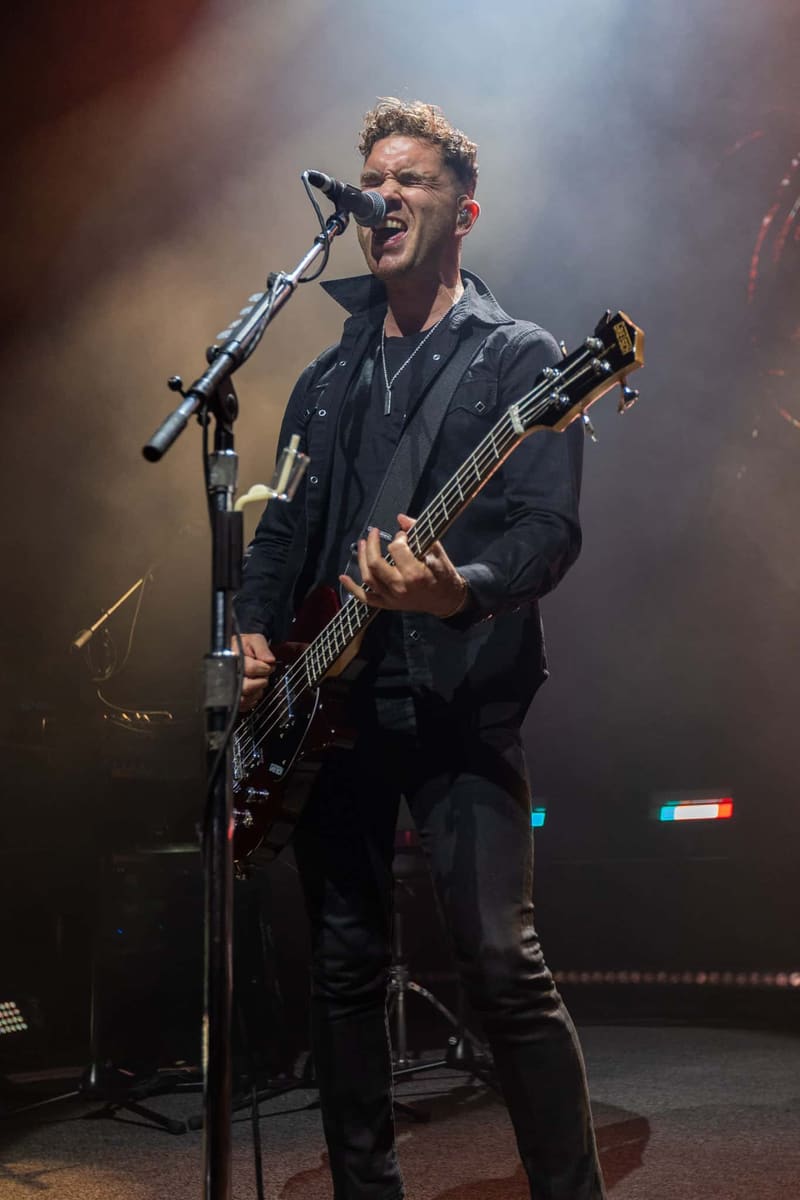 Bassist performs at microphone in dark jacket under warm stage lighting during Royal Blood concert.