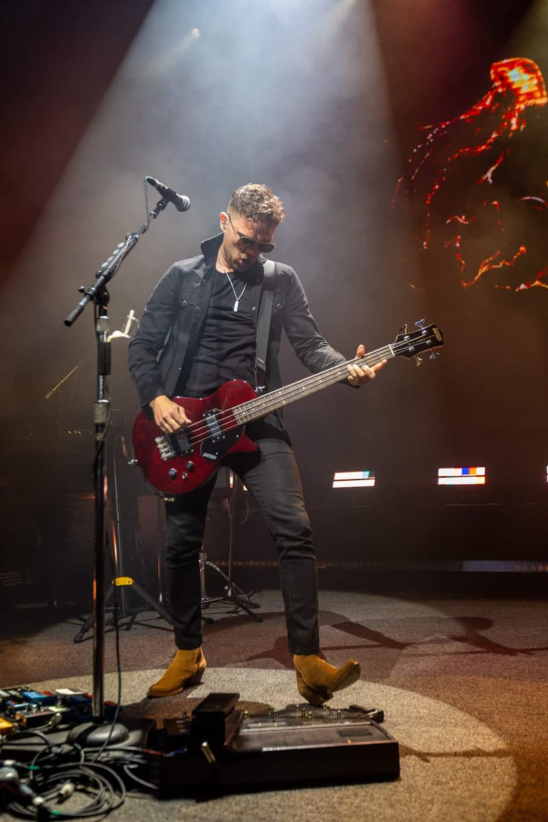 Bassist in dark shirt playing red guitar on stage under dramatic spotlights during Royal Blood performance.