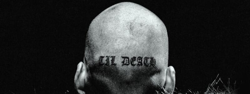 Club Angel - Shaved head with tattooed text viewed from behind against dark background, surrounded by wild grass.